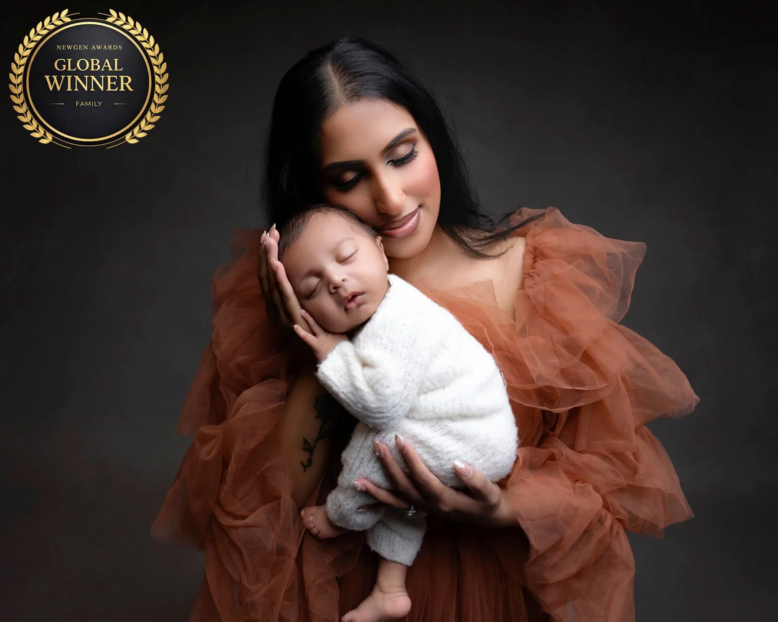 A woman holding a sleeping baby tenderly against her shoulder in a dark studio setting, with a gold laurel award emblem in the top left corner. Auckland newborn photographer