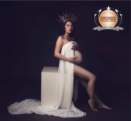 A pregnant woman sitting on a white box, draped in white fabric, with a dark background. She is holding her belly with one hand and has a golden halo-like crown on her head. There is a bronze medal graphic in the top right corner.