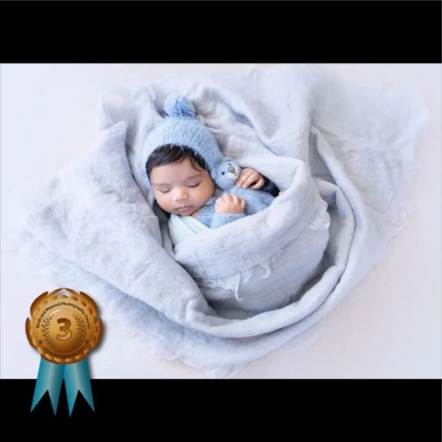 A sleeping baby wrapped in a soft gray blanket, wearing a blue knit hat, with a small badge in the bottom left corner indicating third place award.