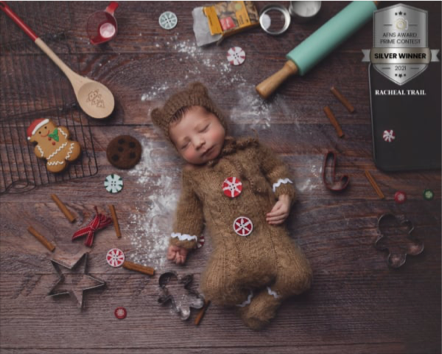 A sleeping baby dressed in a brown onesie with a Santa hat lying on a wooden surface surrounded by holiday baking supplies and decorations.