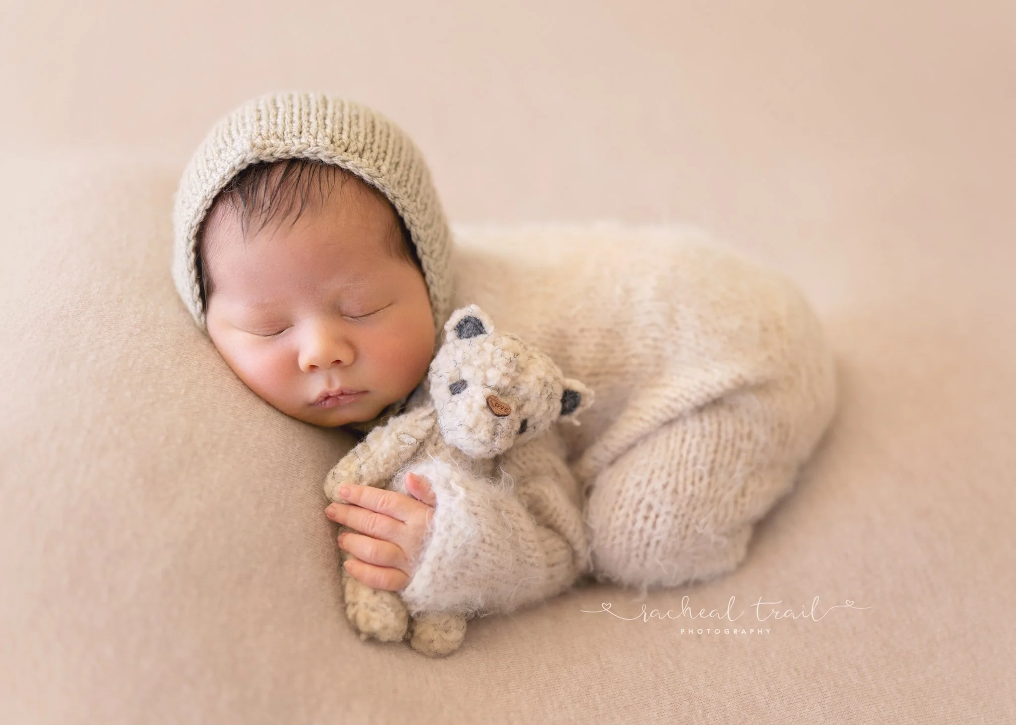 Sleeping baby wearing a knitted hat and sweater, cuddling a small plush teddy bear, on a soft beige surface.