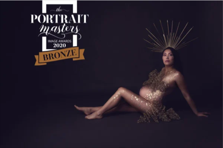 A woman sitting on the floor with a dark background, wearing a golden dress, decorated with gold embellishments, and a crown-like headpiece with radiating gold spikes, in a promotional image for the 2020 Portrait Masters Image Awards Bronze.