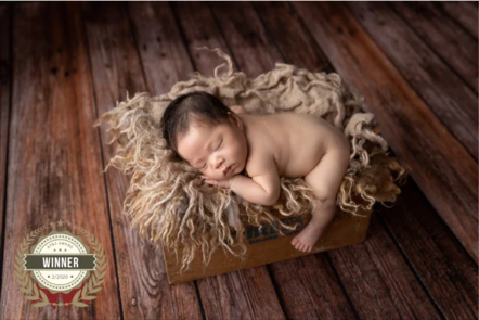 A sleeping baby curled up on a fluffy cream-colored blanket inside a wooden box on a dark wooden floor, with a winner badge in the bottom left corner.