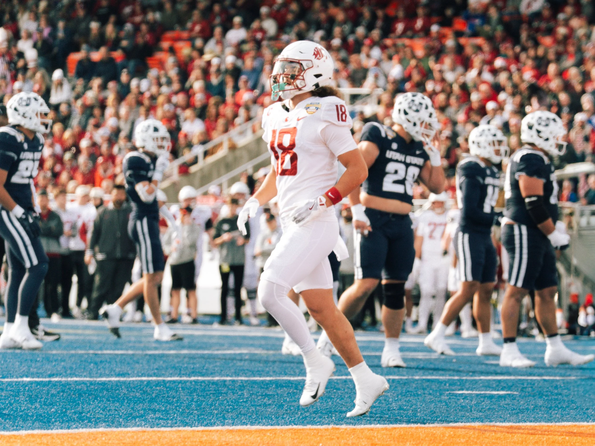 A college football game with players on the field and a large crowd in the stands. A player in white, number 18, is walking with a confident posture, surrounded by players in blue uniforms.