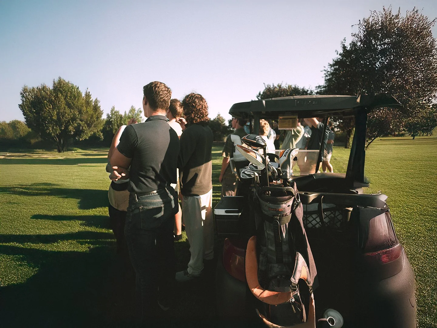 A group of people standing next to a golf cart on a golf course with trees in the background, green grass, and clear skies.