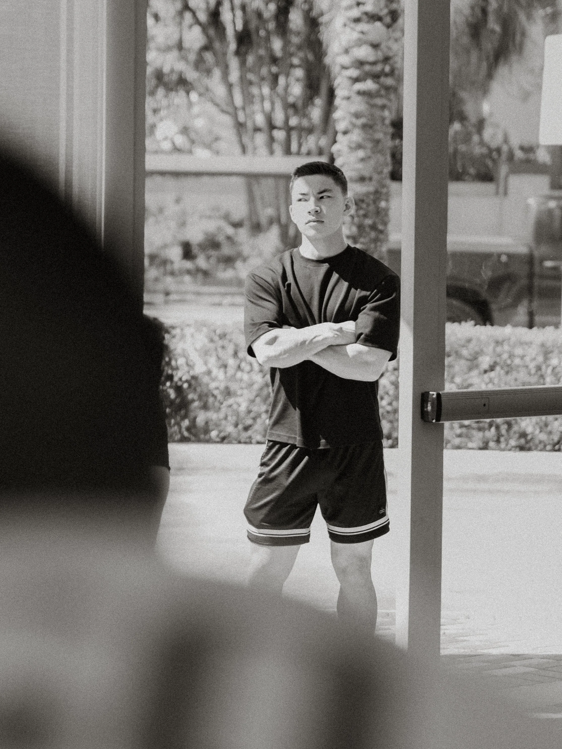 A young man standing outdoors with arms crossed, wearing a black t-shirt and shorts, looking to the side with a serious expression.