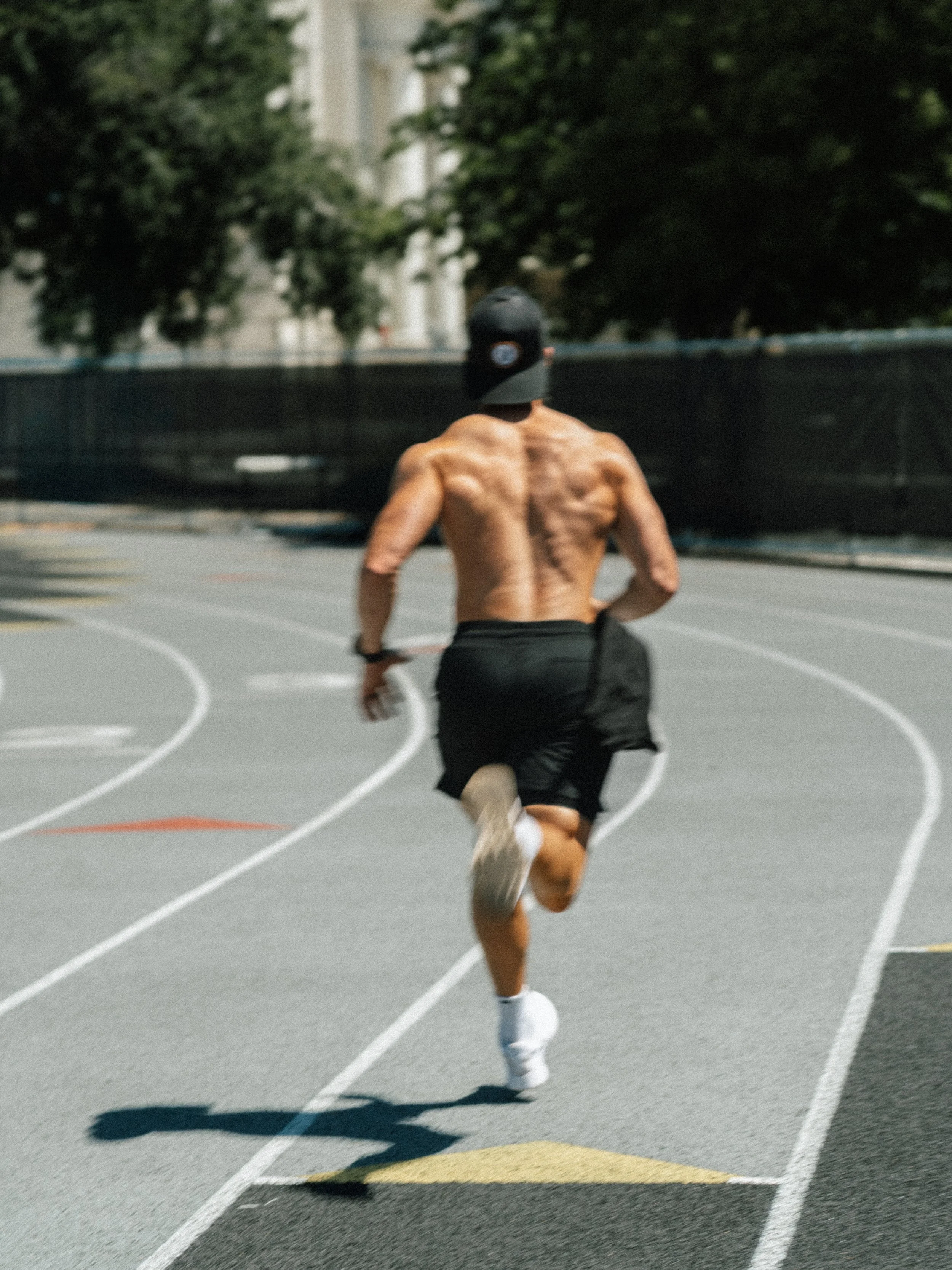 A shirtless man running on an outdoor track wearing black shorts, white shoes, and a black cap, seen from behind.