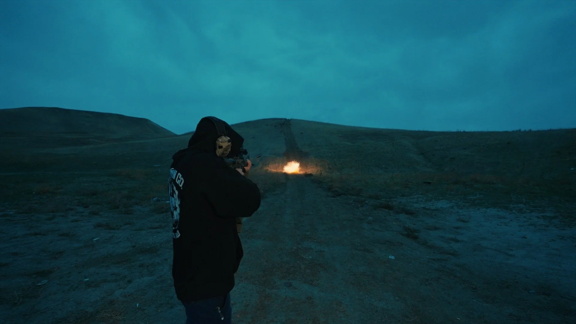 Person in dark clothing aiming a rifle at an explosion in the distance on a dirt path in a hilly, barren landscape at dusk.
