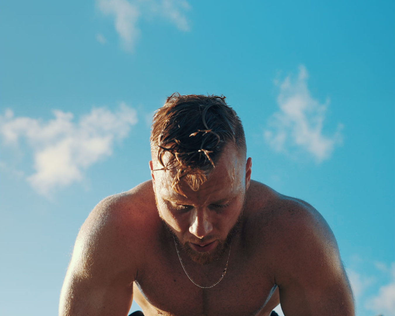 A shirtless man with wet hair and a necklace is looking down, with a blue sky and scattered clouds in the background.