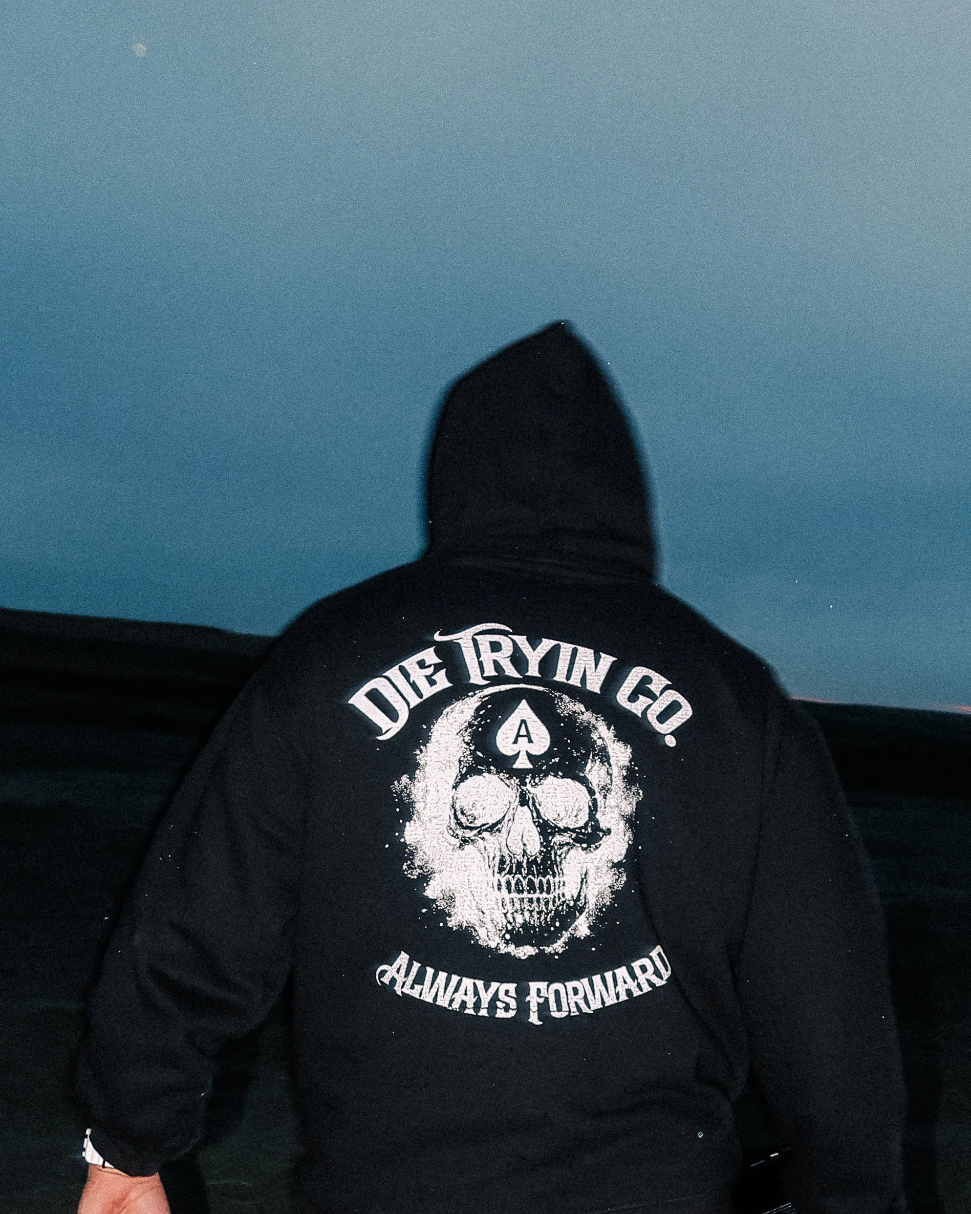 Person wearing a black hoodie with a skull graphic and the words 'DIE TRYIN GO. ALWAYS FORWARD' on the back, standing outdoors at dusk or night.