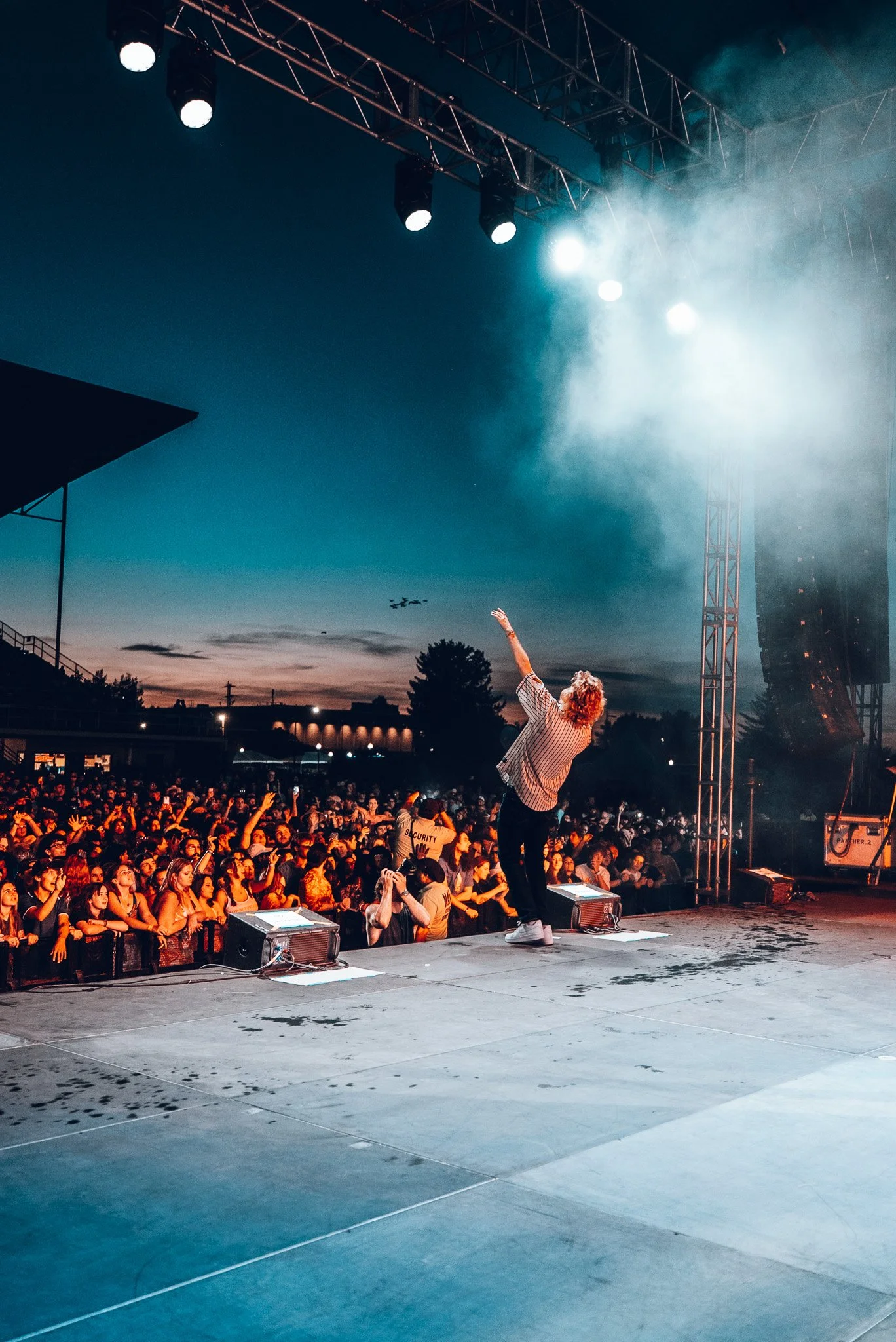 A performer on stage at an outdoor concert during sunset, with a large crowd watching and a sky with clouds and birds.