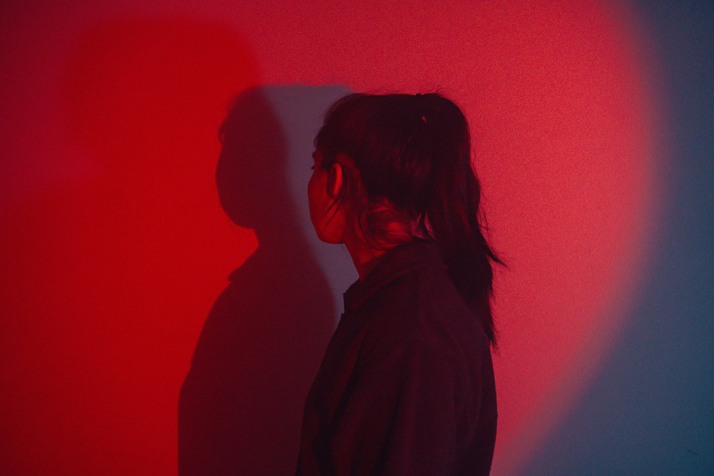 A person with long dark hair stands in front of a wall, casting a shadow that appears as a second person, illuminated by red and dark blue lighting.