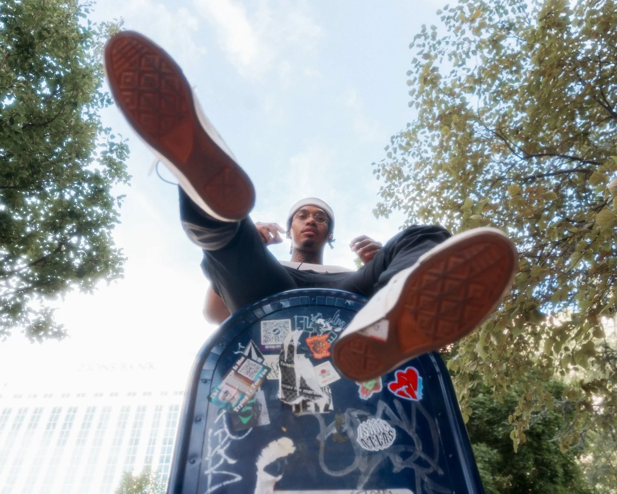 A man wearing glasses and a beanie is seen from below, sitting on a skateboard with a graffiti-covered surface, outdoors among trees, with a city building visible in the background.