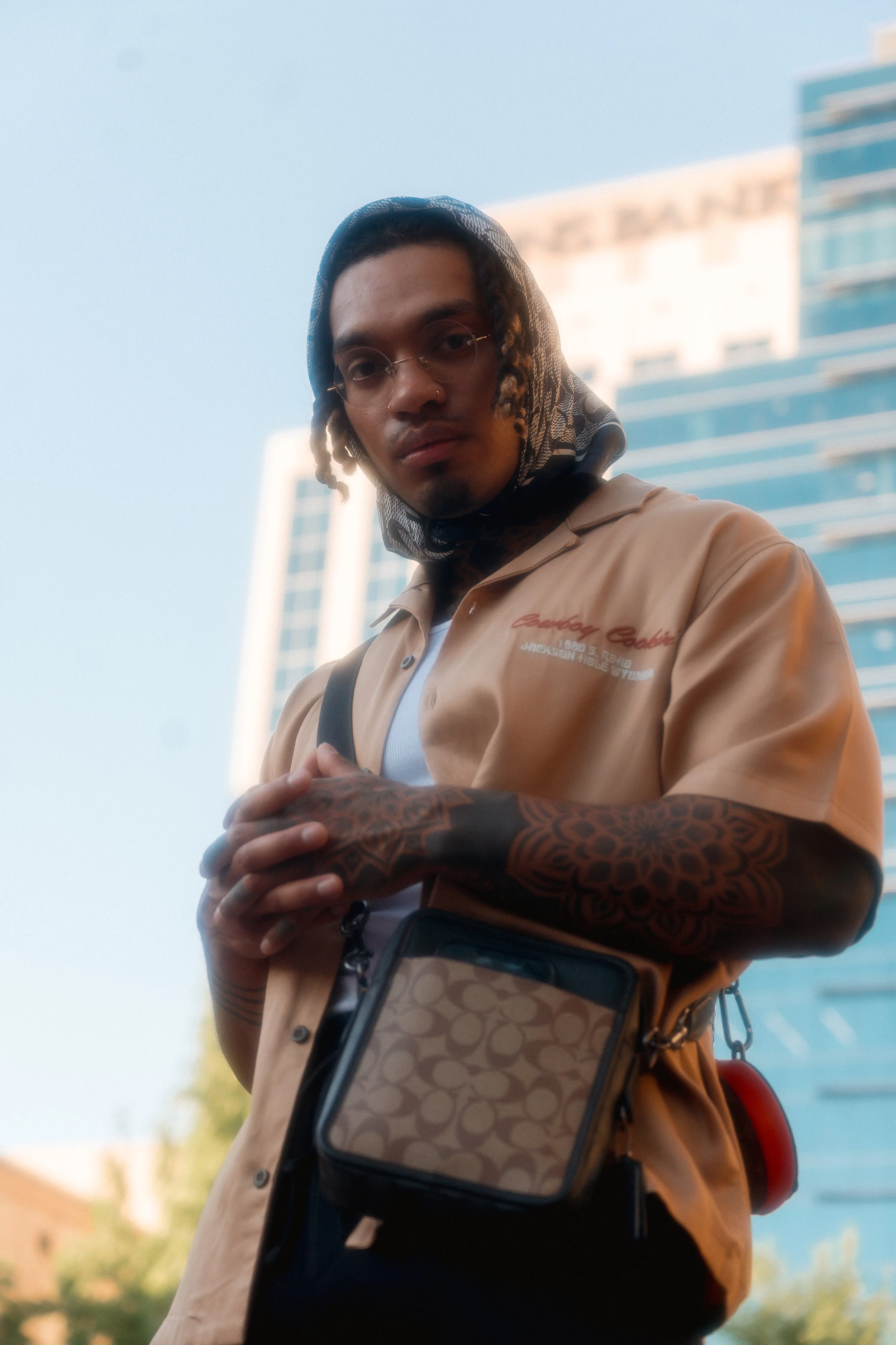 A young man with glasses, dreadlocks, and a scarf covering his head, standing outdoors in front of modern high-rise buildings. He is wearing a beige shirt with red text, has tattoos on his hands and arms, and is holding his hands together in front of him. He also carries a patterned crossbody bag and a small red round object attached to his waist. The sky appears clear and blue.