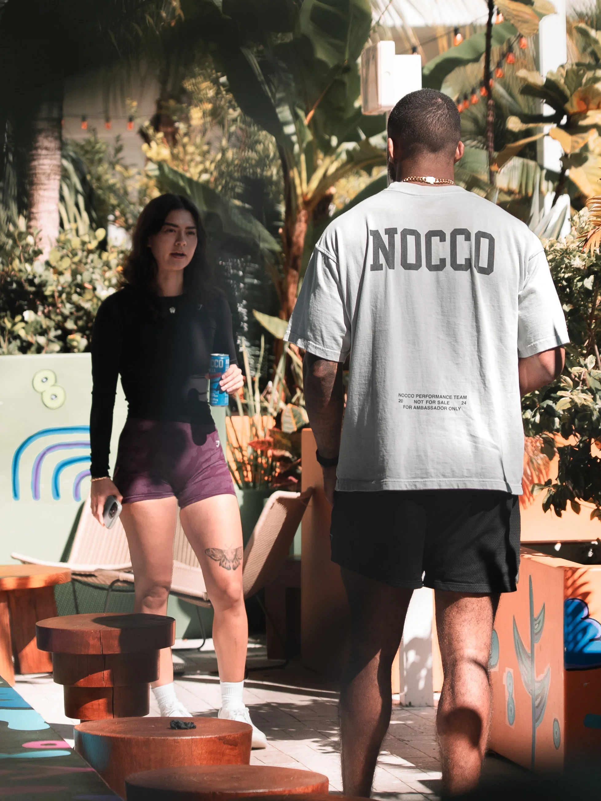Two people talking outdoors among tropical plants. The woman on the left holds a blue drink and a phone, wearing a black top and purple shorts, with tattoos on her thigh. The man on the right has his back to the camera, wearing a white T-shirt with 'NOCCO' on the back and black shorts.