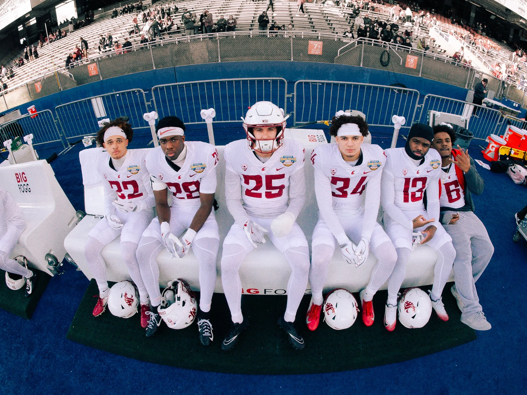 American football players sitting on the team bench in their uniforms at a stadium, with empty seats and spectators in the background.