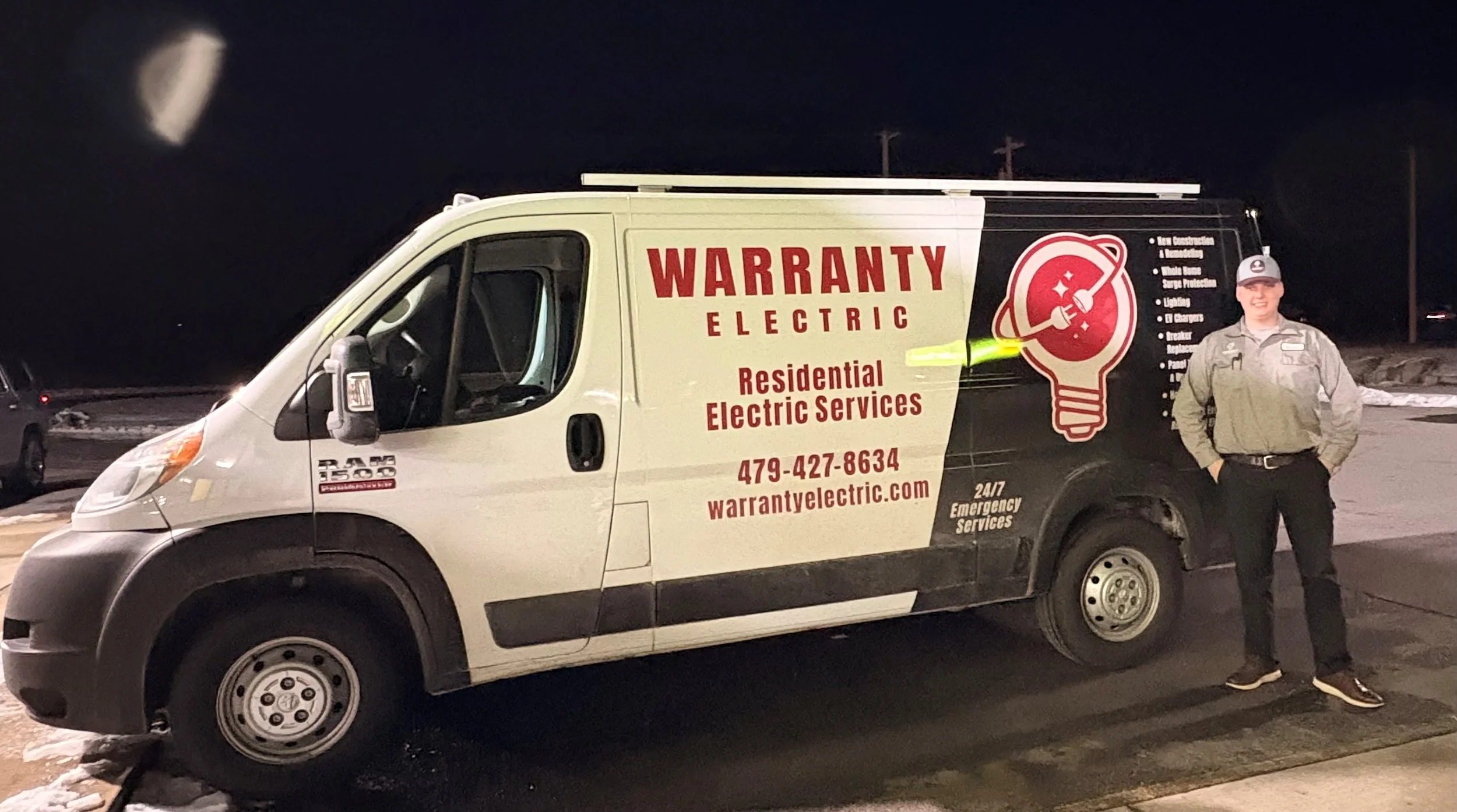 A man standing next to a white and black service van for Warranty Electric at night on a snowy road. The van displays services offered and contact information.