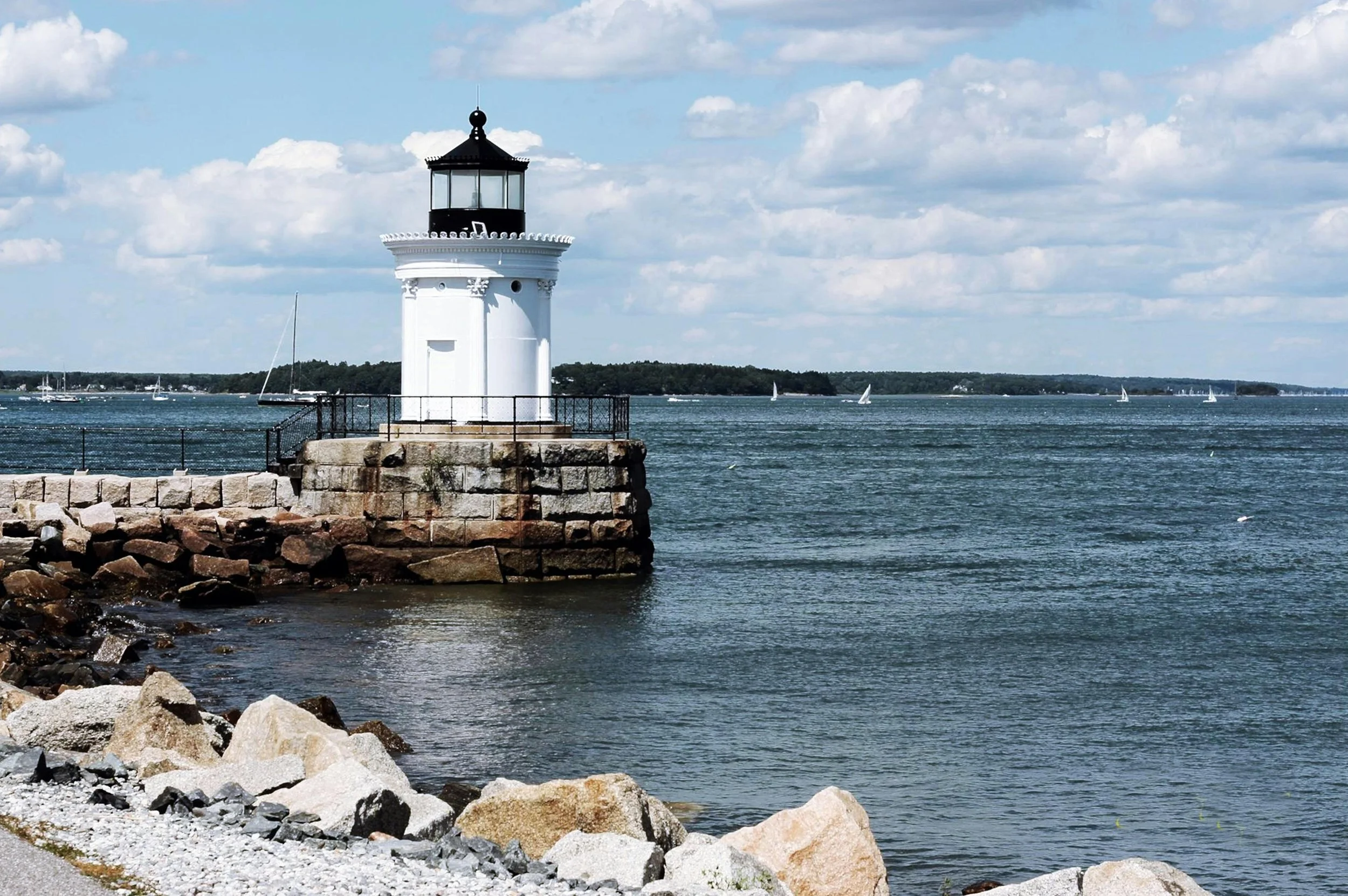 Lighthouse on a rocky breakwater with sailboats on a calm lake under a partly cloudy sky.