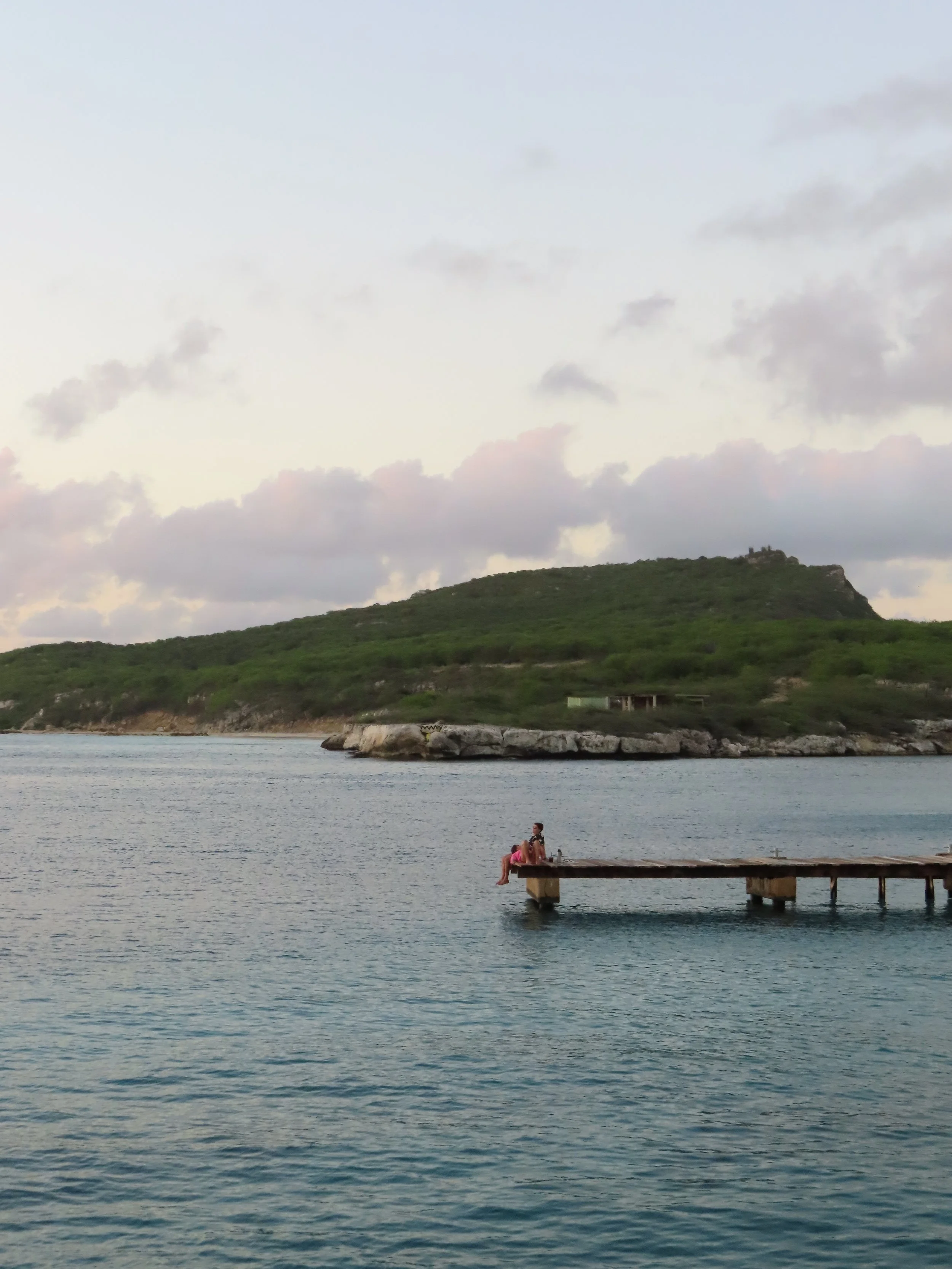 Person sitting on a wooden dock extending into a calm body of water with a green hill and a cloudy sky in the background.