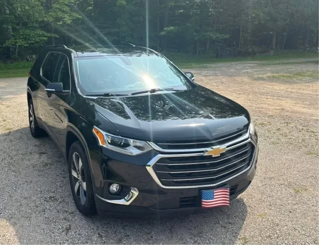 Black Chevrolet Traverse SUV with American flag license plate parked on gravel outdoors, with trees in the background.