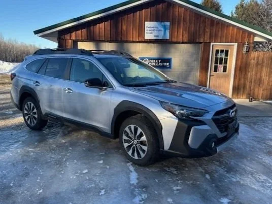 2025 silver Subaru Outback completed repairs parked outside a wooden building with a sign, on a snowy ground.