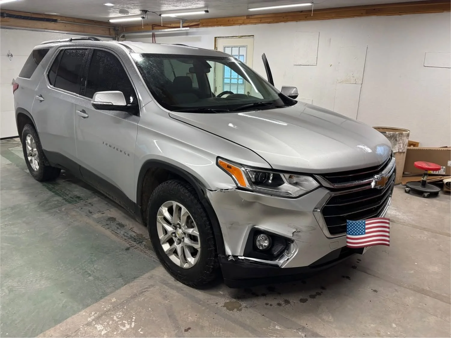 Silver Chevrolet Traverse SUV with front-end damage parked in a garage, American flag covering the license plate.