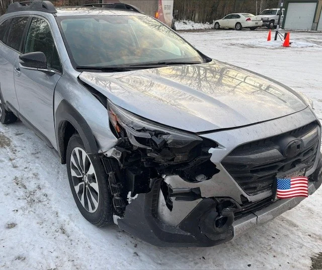 2025 Silver Subaru Outback with significant front-end damage, missing front bumper, and hood partially bent, parked on snow-covered ground.