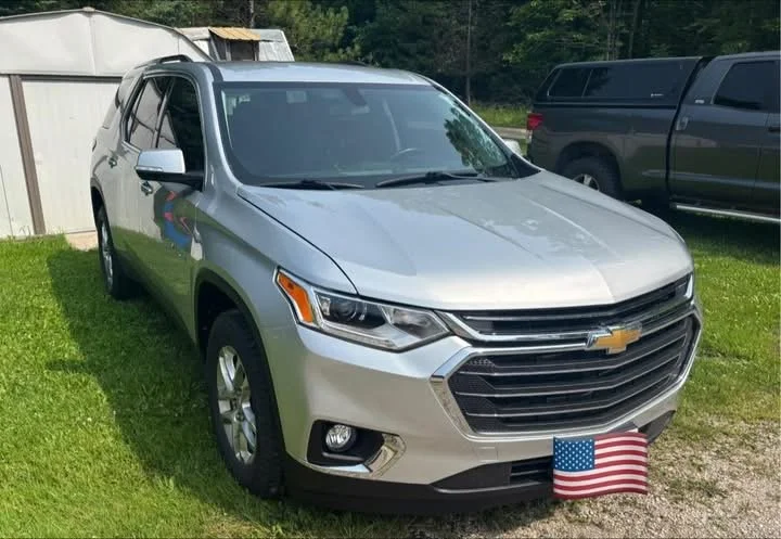 A silver Chevrolet Traverse SUV parked on a grassy area with an American flag positioned in front of the license plate. Other vehicles and a wooden shed are visible in the background.