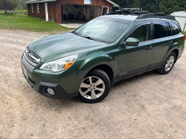 A green Subaru Outback station wagon parked on a gravel driveway outside a barn with open doors.
