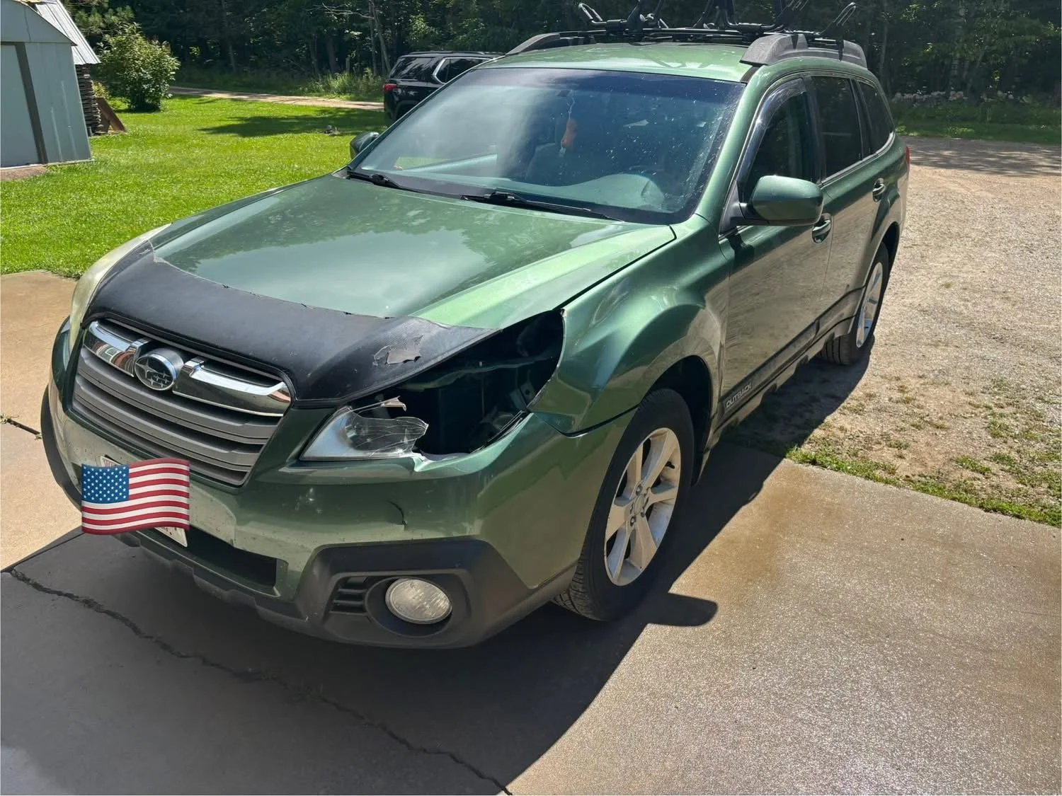 Green Subaru Outback with front-end damage, missing headlight, and a black protective cover on the hood.