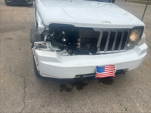 Front of a white Jeep SUV with damage to the front left side and missing headlight, with an American flag hanging from the front bumper.