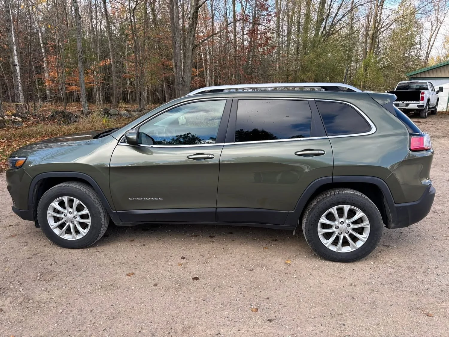 Side view of a green Jeep Cherokee parked on a dirt surface with a wooded area and a white building in the background.