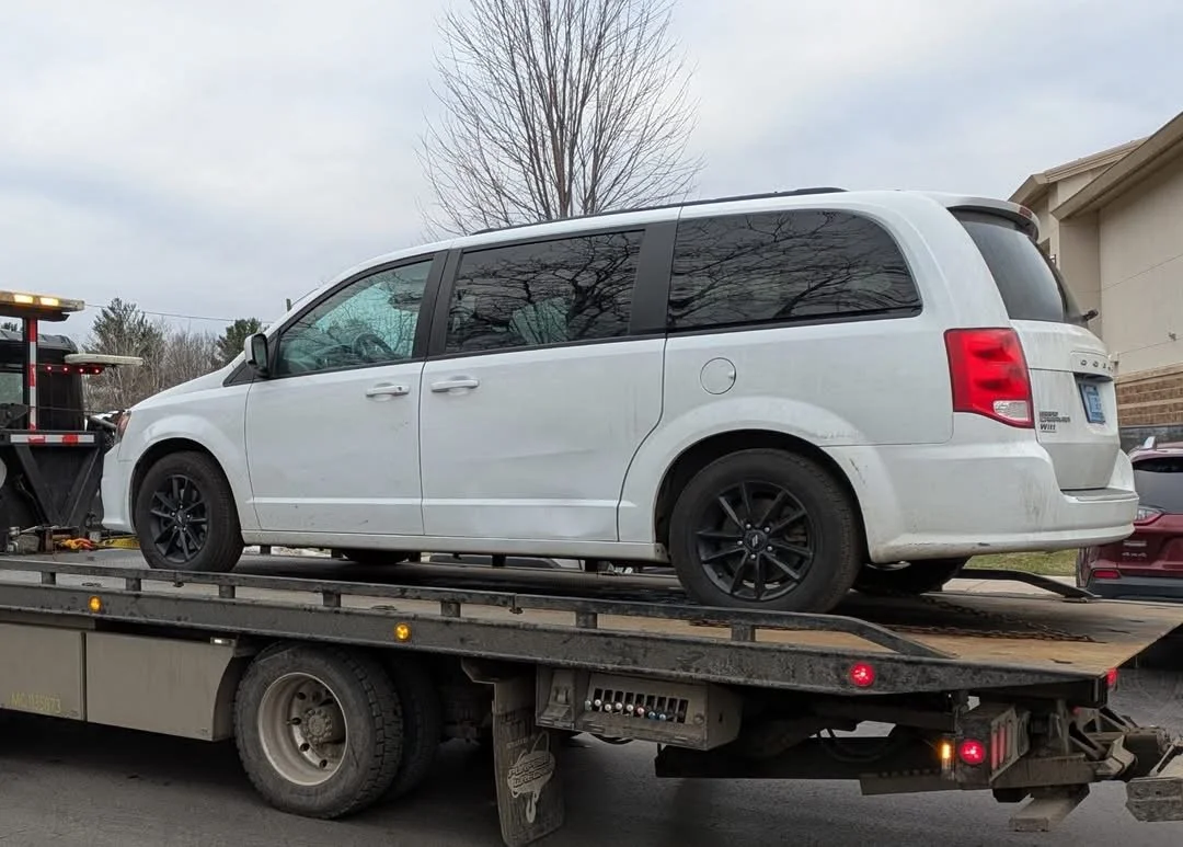 A white Dodge Caravan minivan on a flatbed tow truck, with a leafless tree and house in the background.