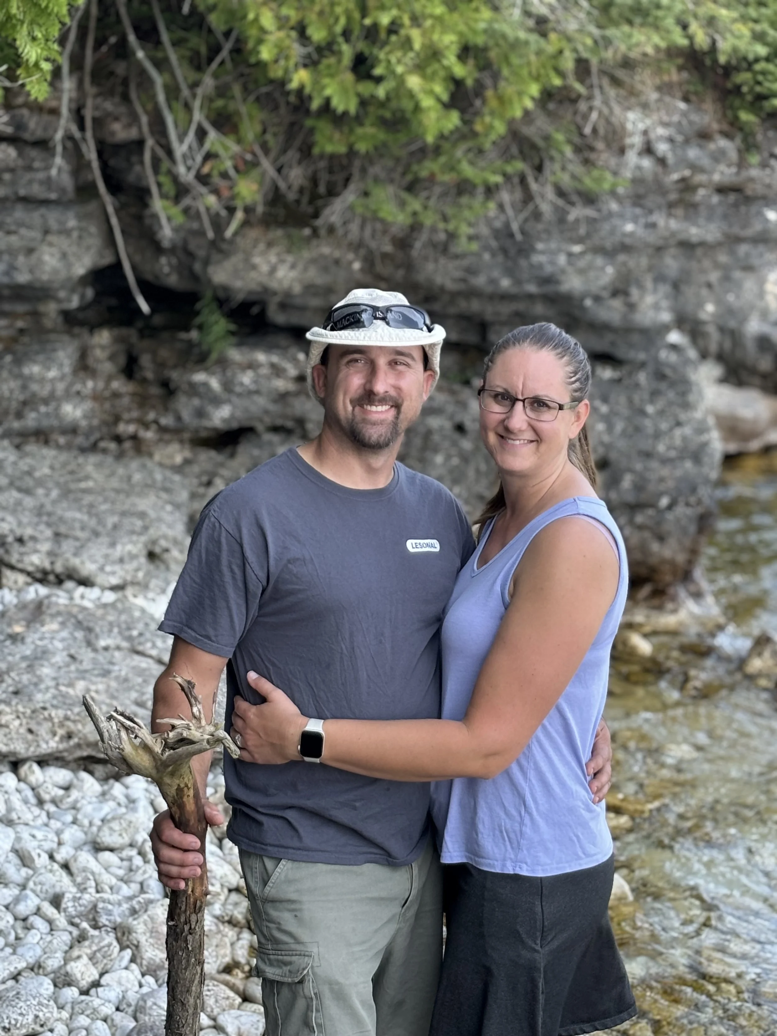 A smiling man and woman standing close together outdoors by a rocky stream, with the man holding a hiking stick and wearing a hat.