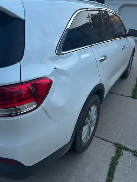 A white KIA Sorento SUV parked on a driveway with a dent on the rear passenger side quarter panel.
