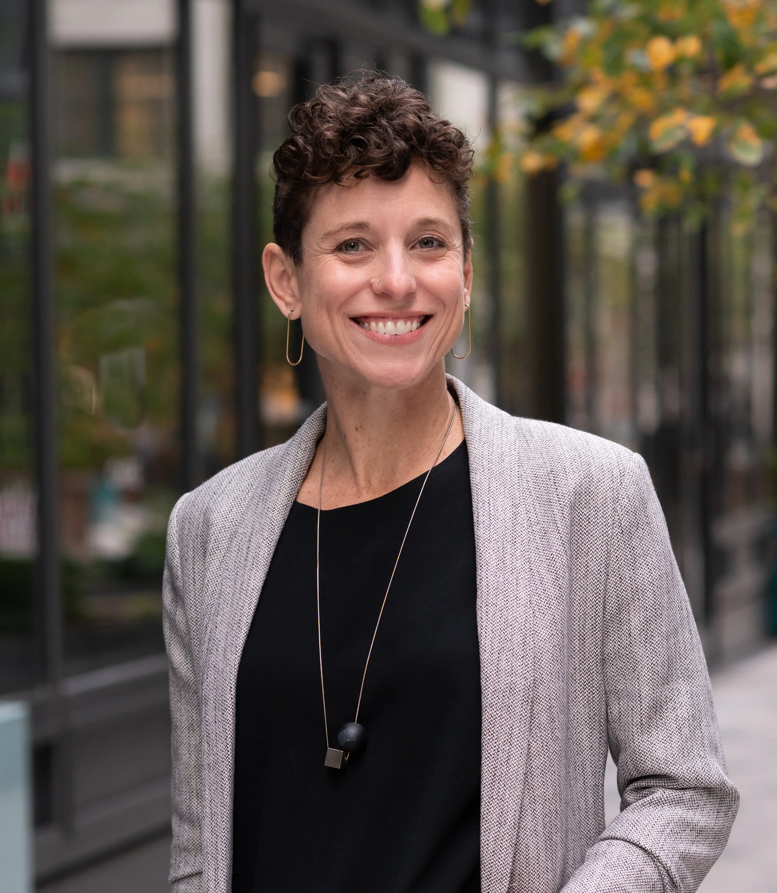 Close-up portrait of a smiling woman with short curly hair, wearing earrings, a black top, and a gray blazer, outdoors with autumn-colored foliage in the background.