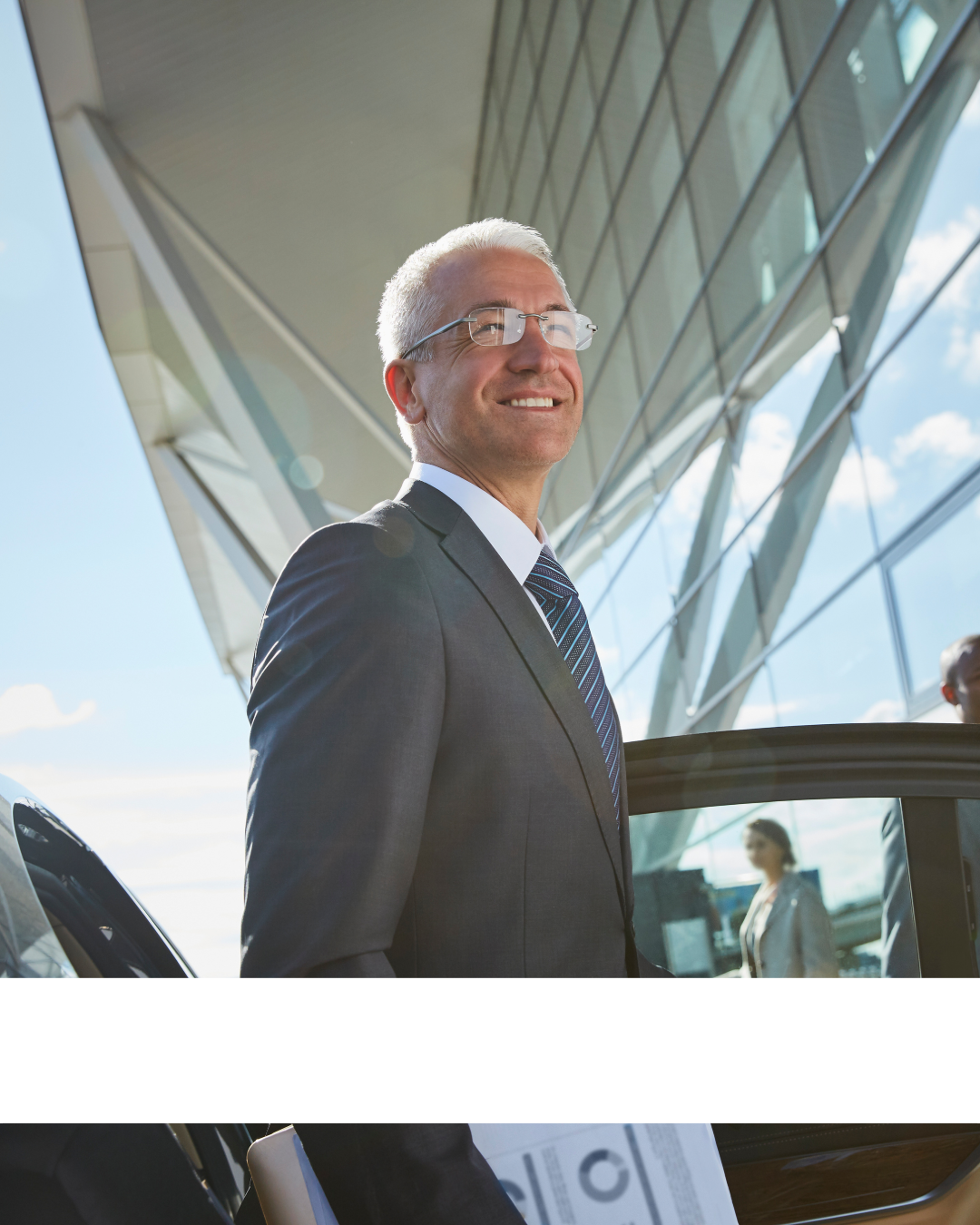 A smiling middle-aged man with gray hair and glasses wearing a dark suit and striped tie standing outside a modern glass building.