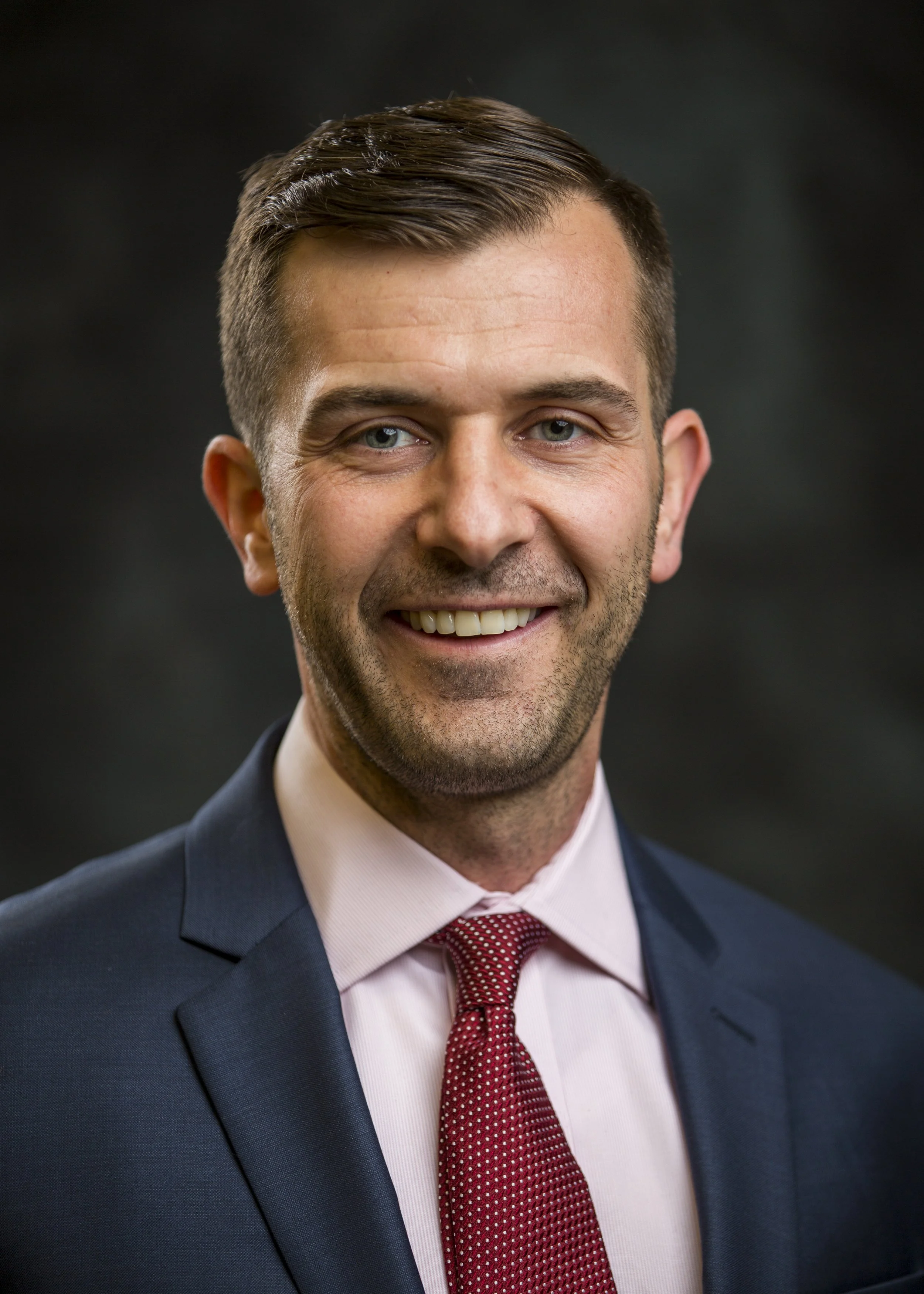 Close-up portrait of a smiling man in a suit and red tie, with short, neatly styled hair against a dark background.