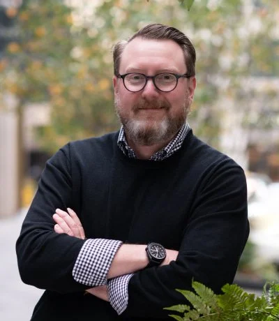 A man with glasses and a beard smiling outdoors, wearing a black sweater and checkered shirt, with autumn trees and buildings in the background.