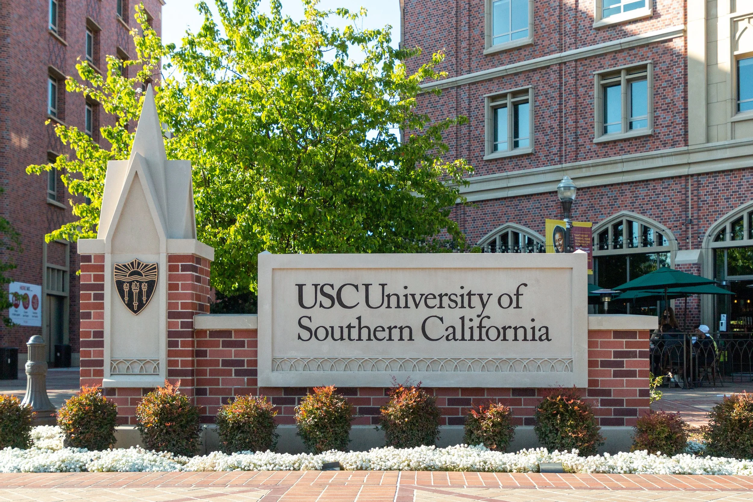 Entrance sign for USC University of Southern California in front of red brick campus buildings, with green trees and flower beds.