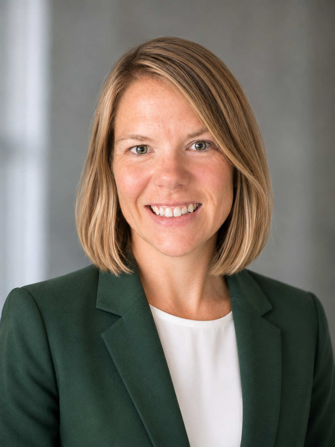 A professional woman with shoulder-length blonde hair, wearing a green blazer and white blouse, smiling at the camera.