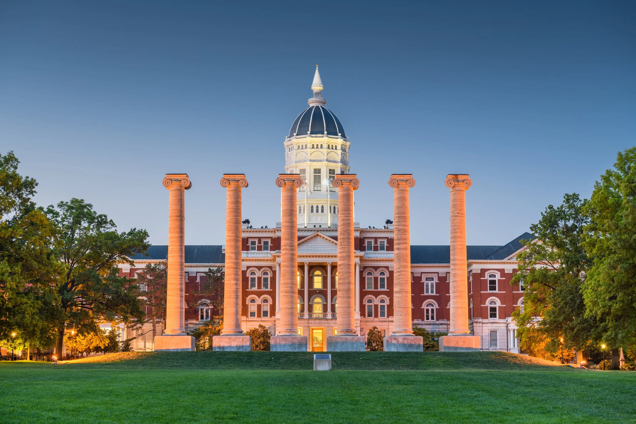 Historic building with a large dome, tall columns in front, and lush green trees and grass surrounding it during the evening.