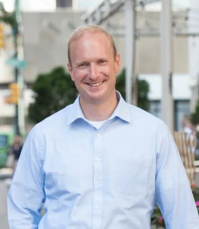 A smiling man with short hair wearing a light blue button-up shirt, standing outdoors in an urban setting.