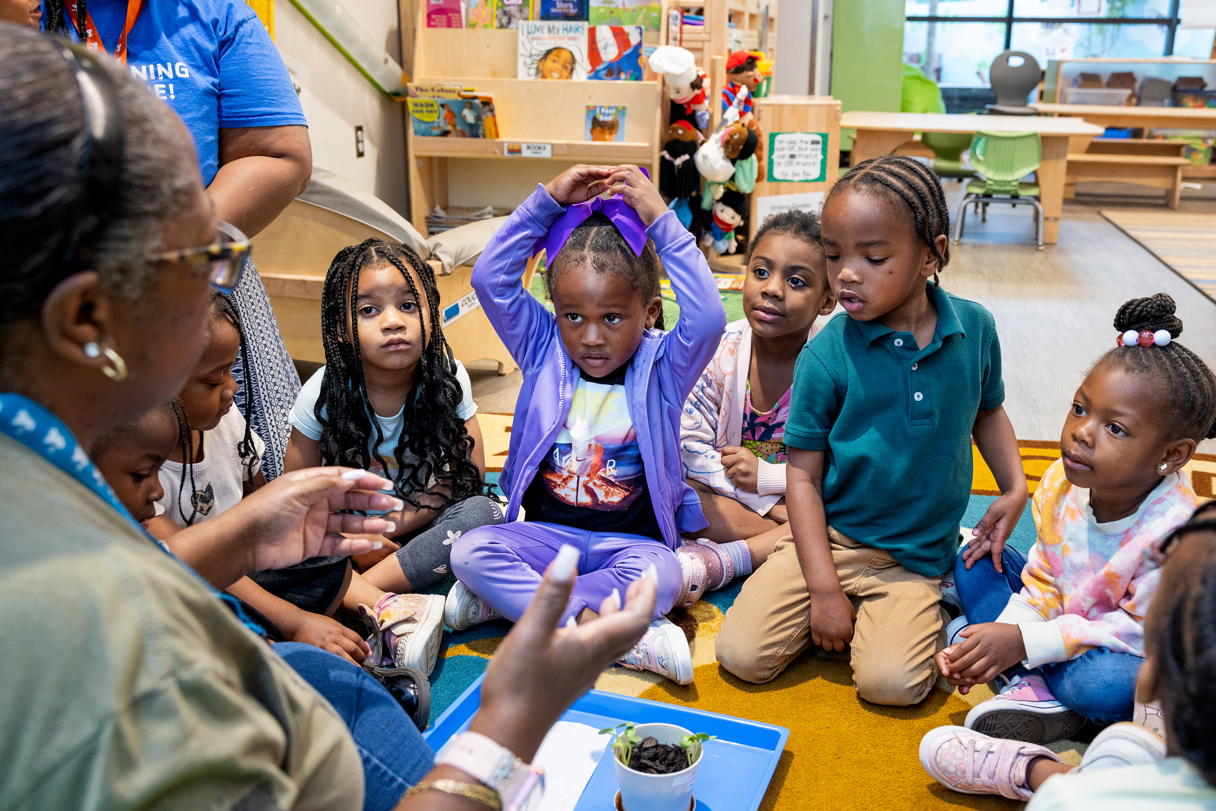 A group of young children sitting on the floor in a classroom, listening to an adult. One girl in the center is raising her hand, surrounded by books, toys, and shelves in the background.