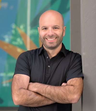 A smiling man with a shaved head and beard, wearing a black polo shirt, standing with arms crossed in front of a colorful, abstract background.