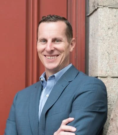 Professional man in a blue suit leaning against a stone wall with a red wooden door in the background.