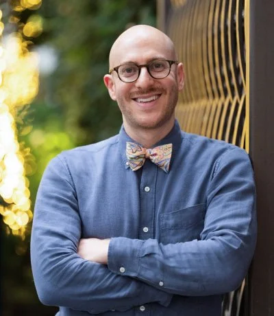 A smiling man with glasses, a bow tie, and a blue button-up shirt standing outdoors next to a wooden fence.