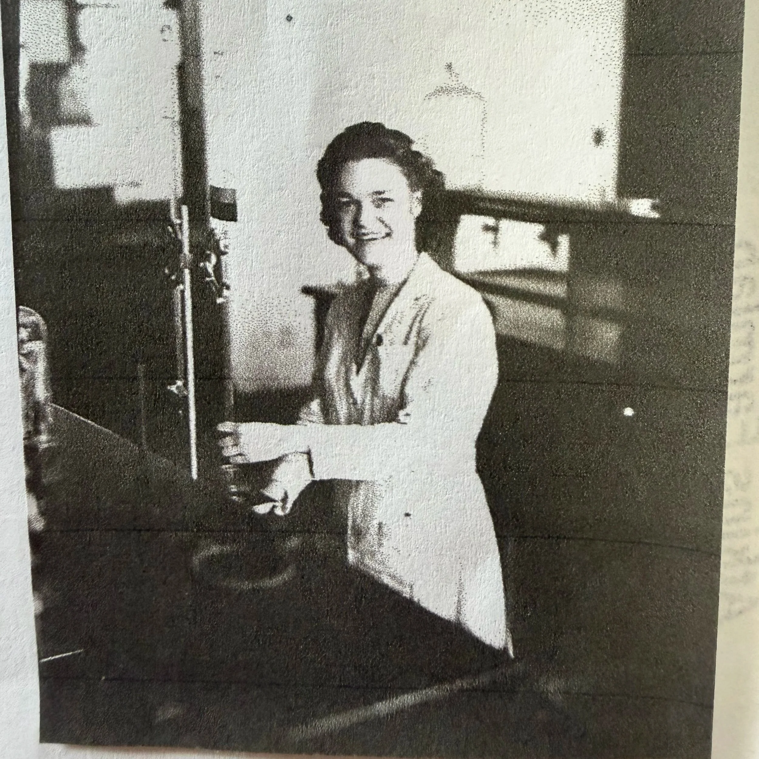 Black and white photo of a woman sitting at a piano, smiling and looking at the camera.