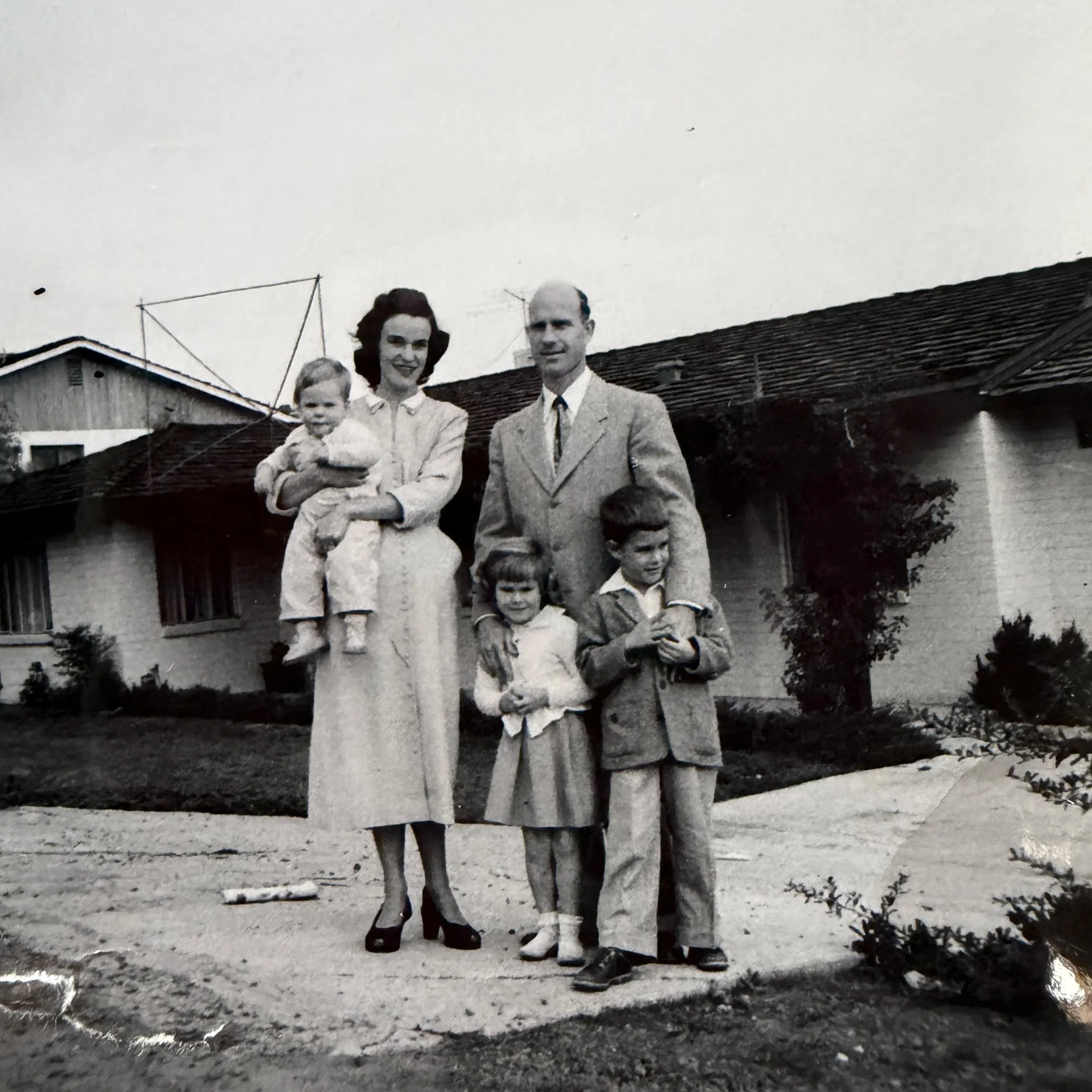 A black-and-white photo of a family standing outside a house, including a woman holding a small child, a man, and two children standing in front of them.