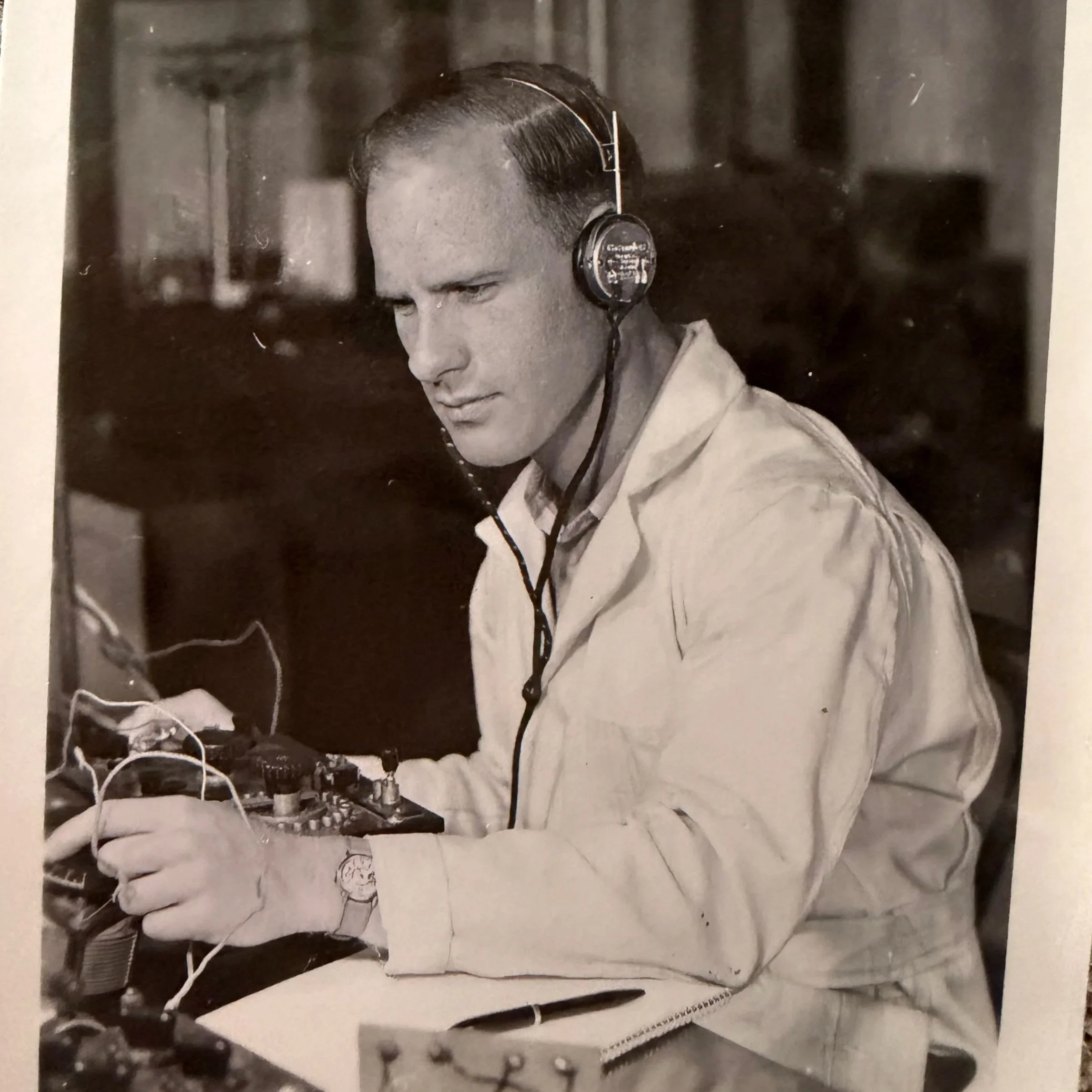 A man in a work coat and wristwatch wearing vintage over-ear headphones, working with electronic equipment or audio devices at a desk.
